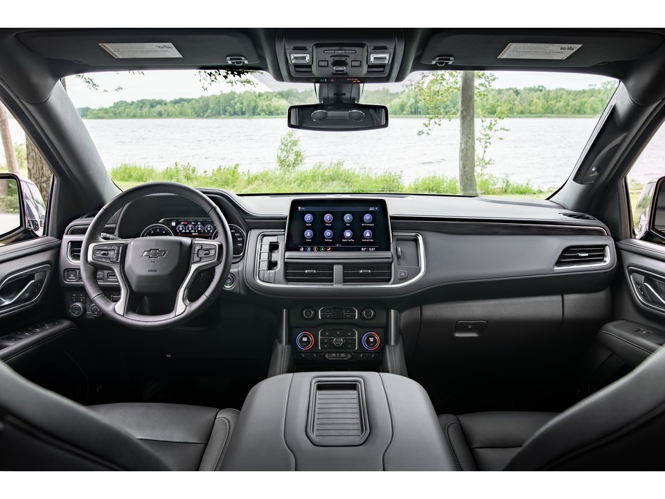 Interior view of a Chevrolet Suburban, showcasing the dashboard, steering wheel, and center console with a large touchscreen display, set against a scenic background of a lake and trees.